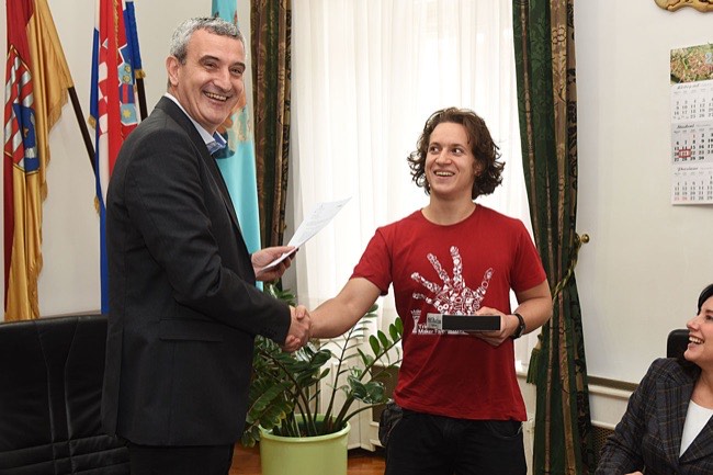 Albert Gajšak shaking hands with an official and receiving a certificate in a formal room with Croatian flags.