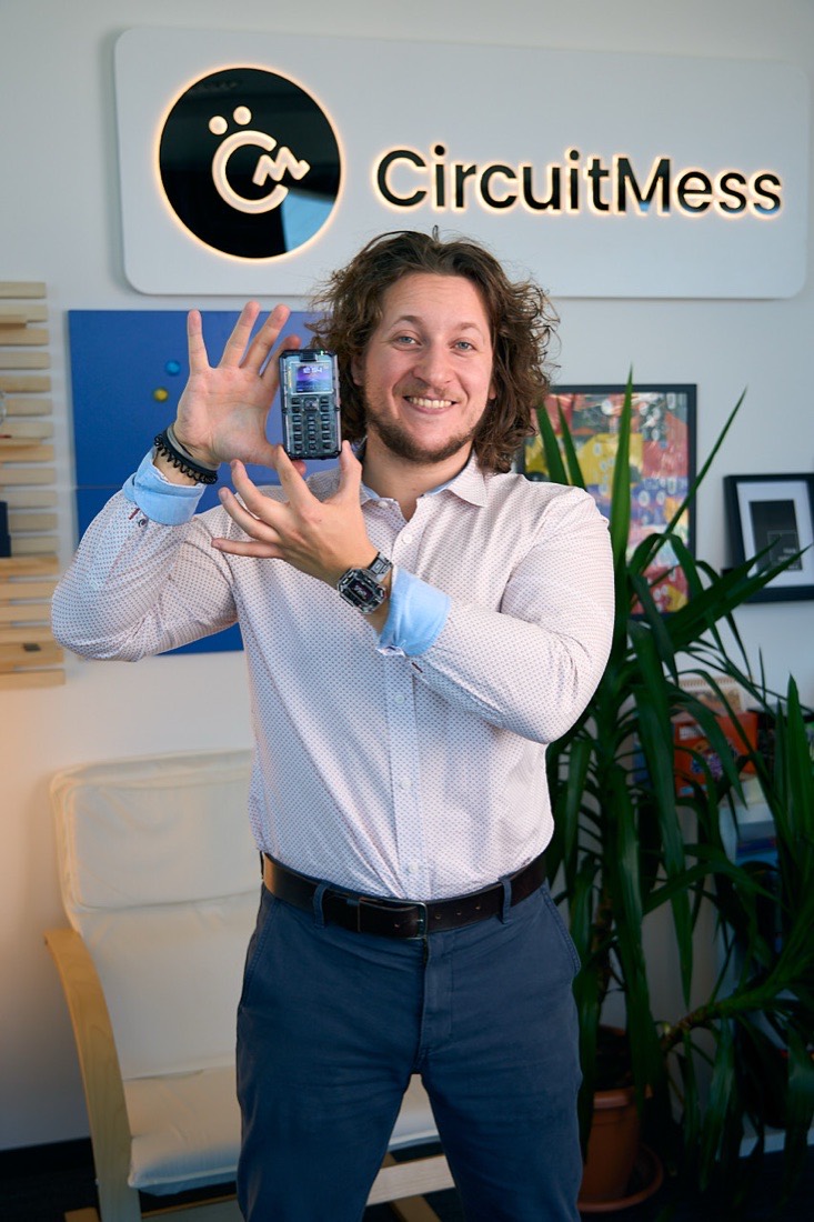 Albert Gajšak standing in the CircuitMess office, holding up a MAKERphone kit, with the CircuitMess logo on the wall behind him.