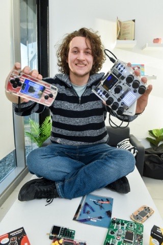 Albert Gajšak sitting cross-legged on a table surrounded by CircuitMess kits, holding one in each hand.