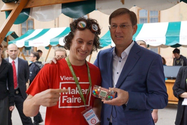 Albert Gajšak in a Vienna Maker Faire T-shirt and goggles, showing a MAKERbuino to a visitor in a suit.