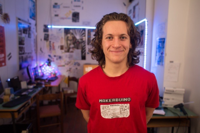 Albert Gajšak in a MAKERbuino T-shirt standing in his bedroom workshop, lit by LED strips.