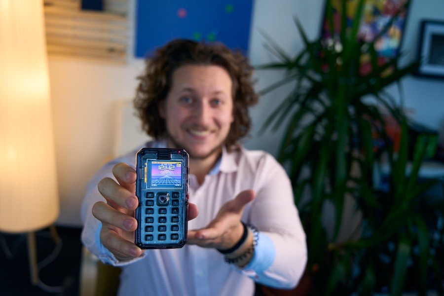 Close-up of a MAKERphone held out towards the camera, its colour screen lit, Albert Gajšak smiling behind it.