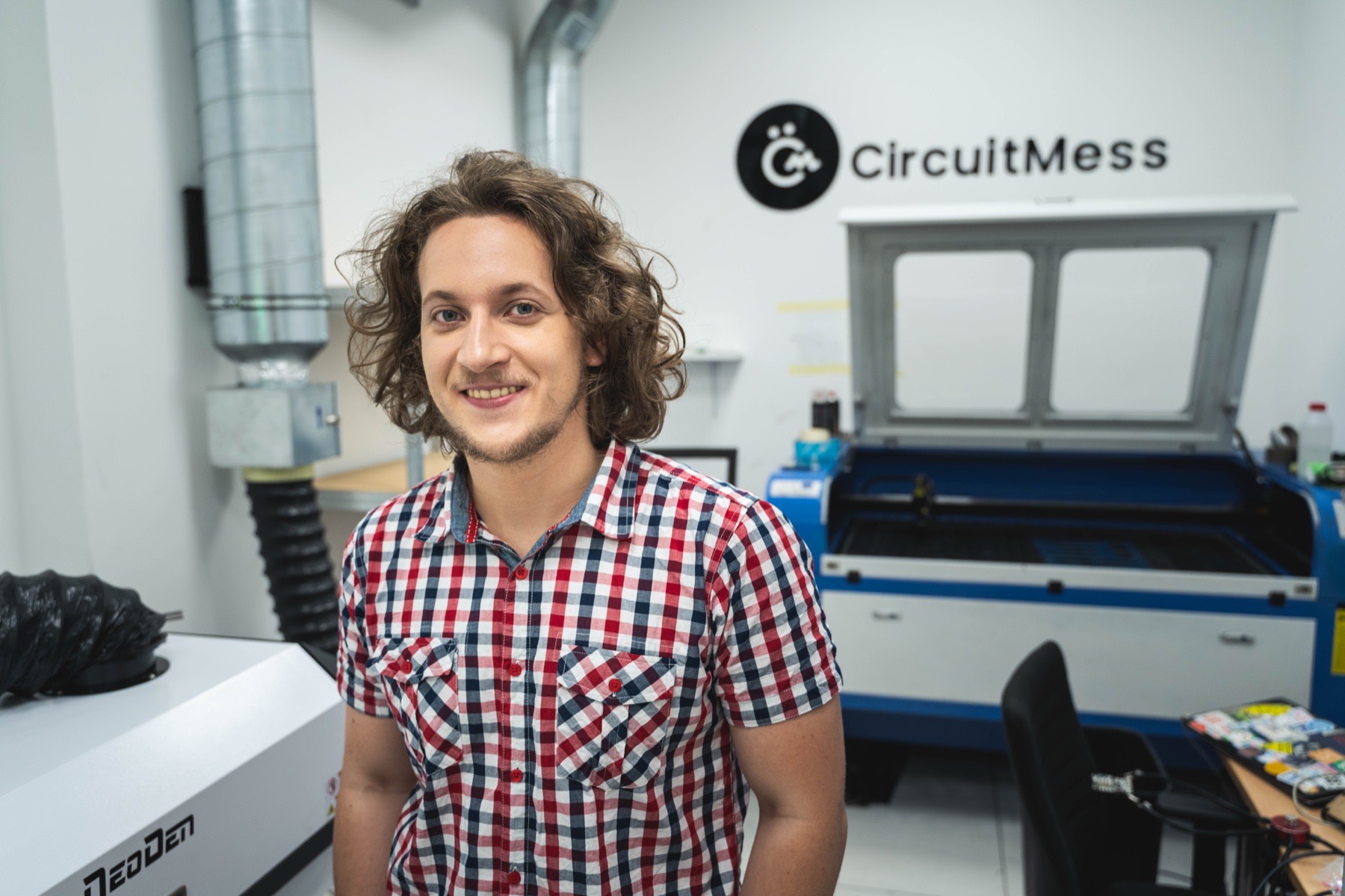 Albert Gajšak in the CircuitMess workshop, standing between a pick-and-place machine and a laser cutter, with the CircuitMess logo on the wall.